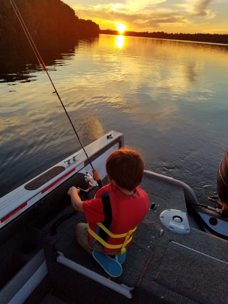 Young boy fishing from boat at golden sunset on lake
